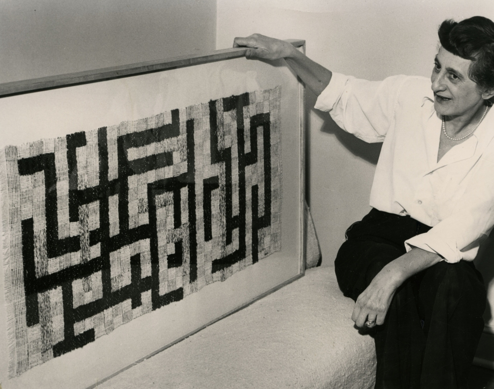 Foto einer älteren Frau, die neben einem geometrischen Textilkunstwerk sitzt.